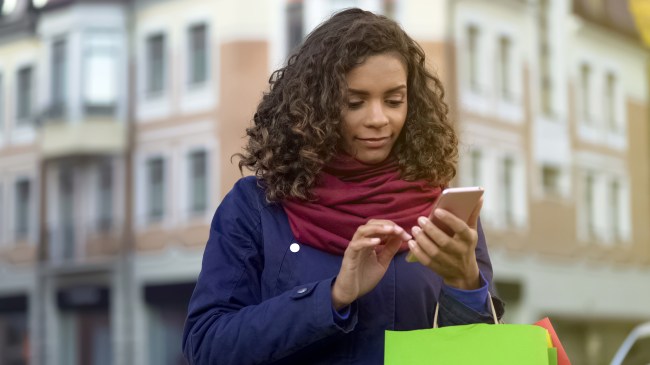 Woman looking shopping on her mobile phone, carrying a few shopping bags. Strong Customer Authentication (SCA) will require a second form of authenticating online purchases. 